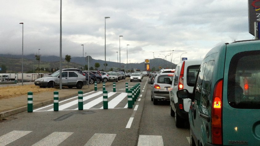 Cua per accedir als aparcaments. Alguns cotxes aparcats a la zona verda.